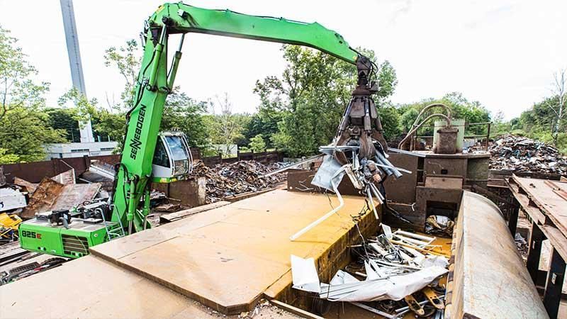 Ein grüner Kran hebt auf einem Schrottplatz einen Stapel Metall an.