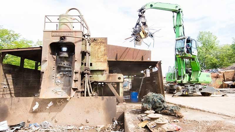 Ein grüner Kran hebt auf einem Schrottplatz ein großes Stück Metall an.