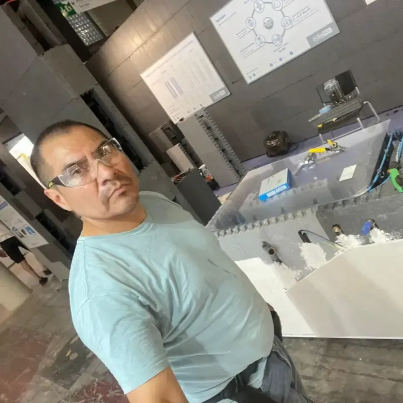 Hombre con camisa azul claro de pie junto a una maqueta de un edificio en una habitación que parece un taller.