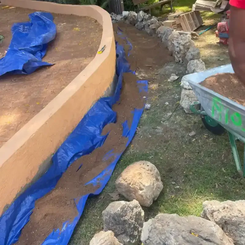 Sendero de jardín en construcción junto a una jardinera de piedra curva, con una lona azul y rocas bordeando el perímetro.