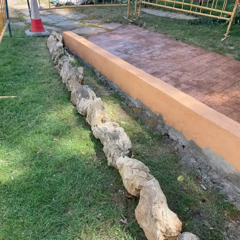 Rampa de hormigón revestida de piedra junto a un sendero cubierto de hierba, con bordillo naranja y pasarela de baldosas.