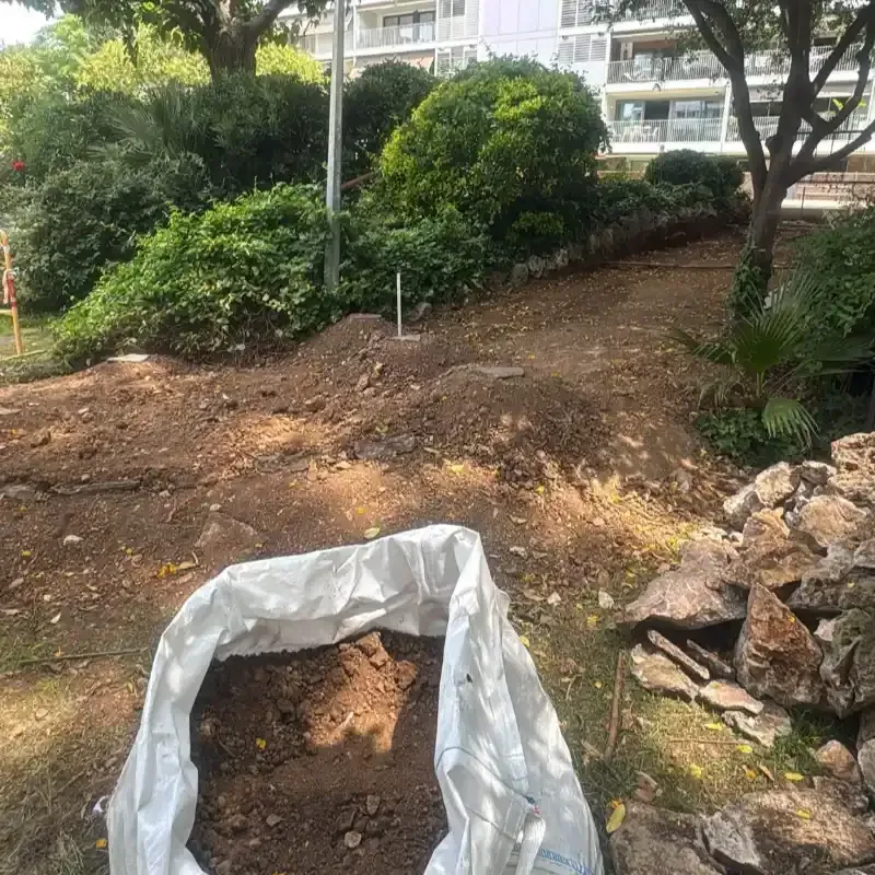 Bolsa blanca de tierra en una ladera ajardinada junto a rocas y arbustos verdes, con edificios de apartamentos arriba.