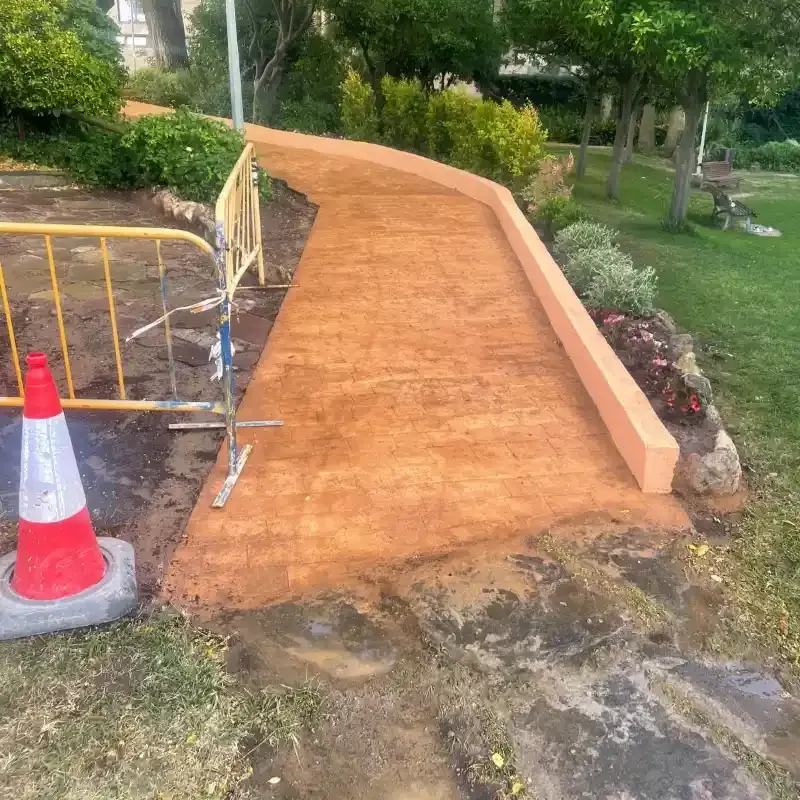 Sendero de color marrón anaranjado recién pavimentado con una rampa junto a un parque con césped, delimitado por un bordillo.