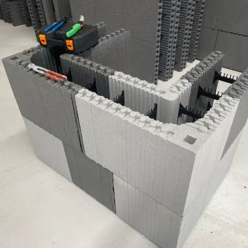 Paredes de ladrillo gris estilo LEGO que forman una pequeña escena interior con dos vehículos de juguete encima y dentro.