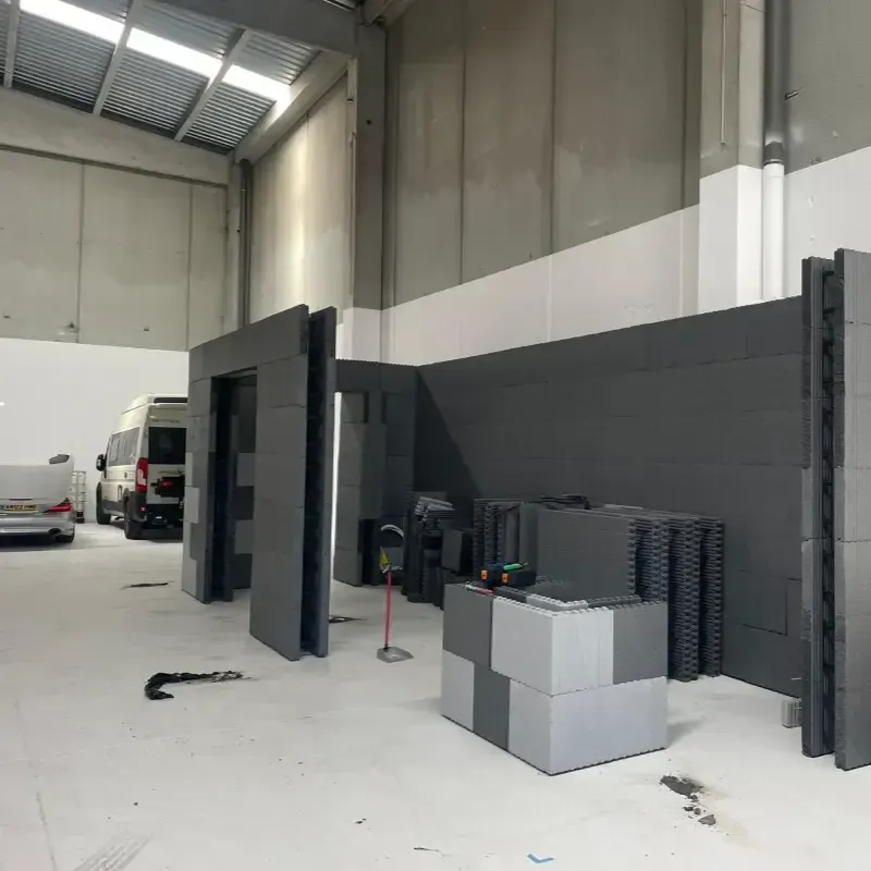 Interior de un almacén industrial con furgonetas aparcadas, baldosas apiladas y una pared gris bajo techos altos.