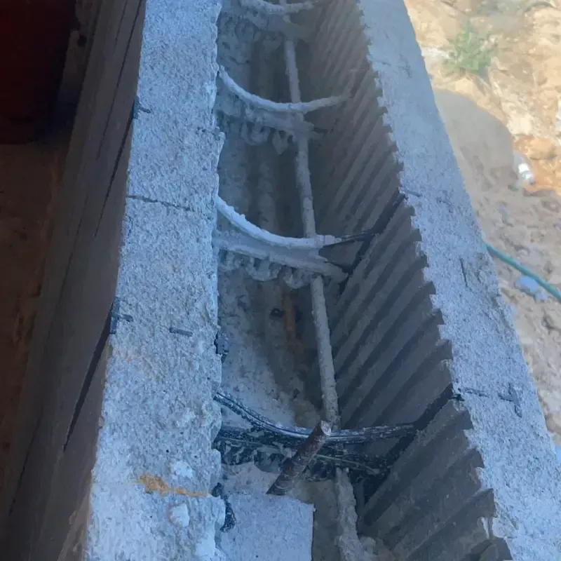 Muro de hormigón dañado con barras de metal expuestas y escombros en una zanja estrecha.