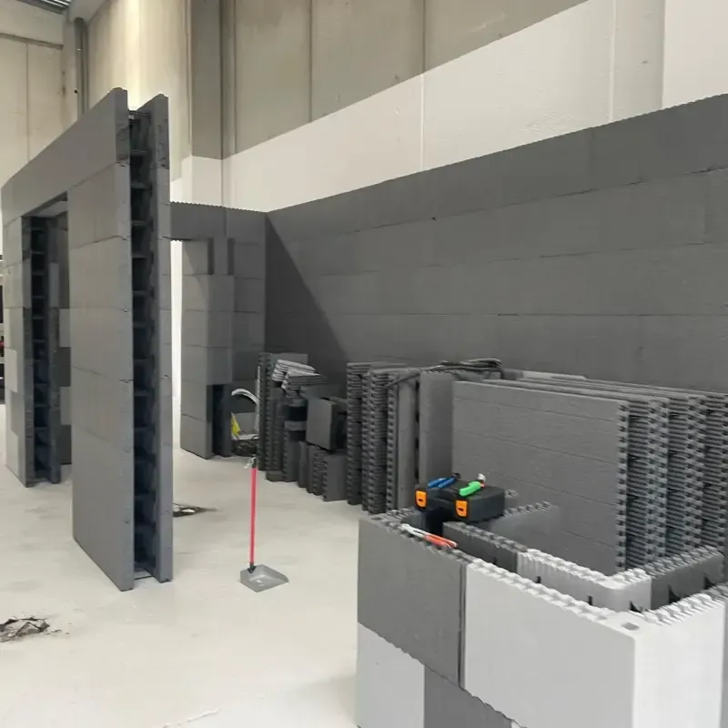 Obra en construcción interior con paredes modulares grises, paneles apilados y un poste de topografía rojo.