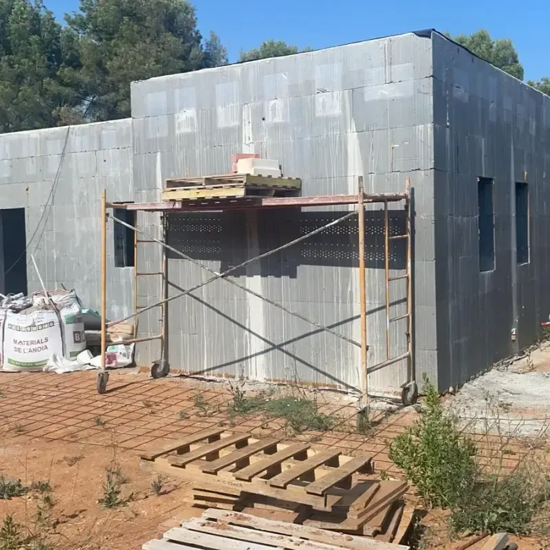 Edificio de metal gris en construcción con andamios, paletas y tierra en primer plano.