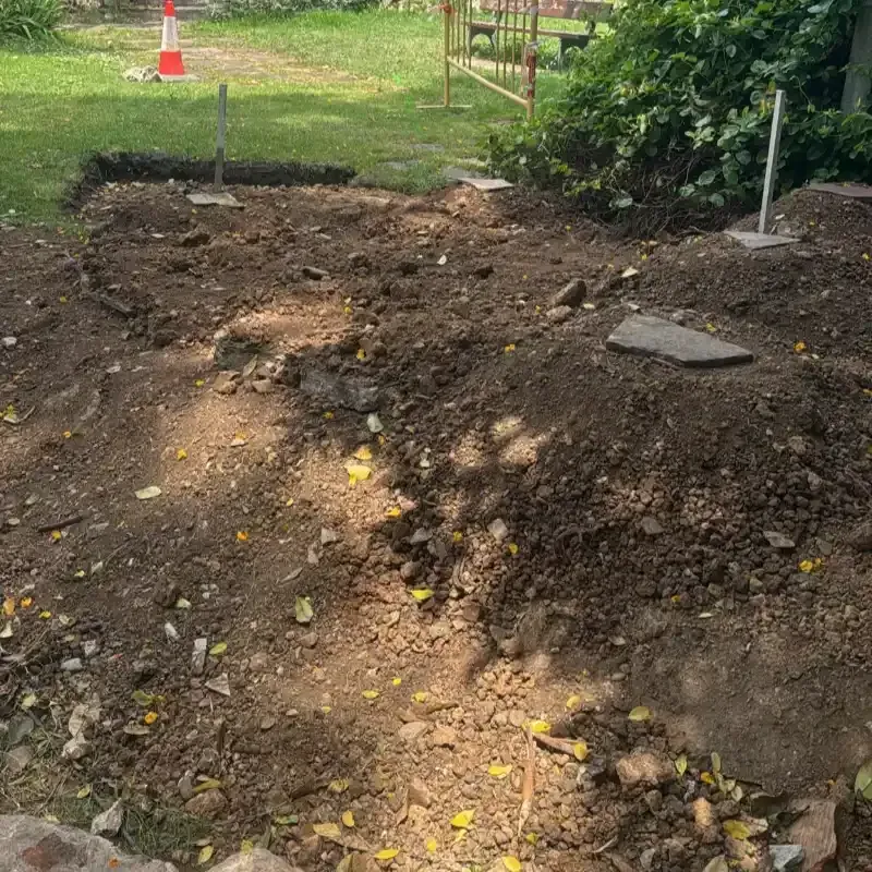 Montón de tierra oscura recién removida en un patio, con pequeñas estacas y un cono al fondo.