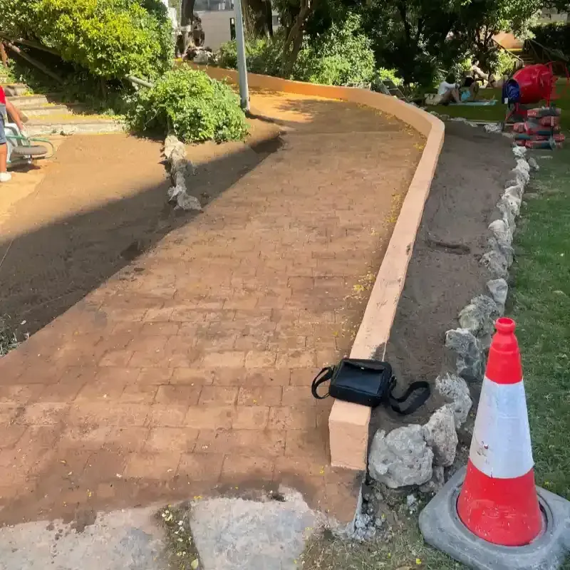 Sendero curvo de ladrillos en construcción, bordeado de rocas y un cono de tráfico en un jardín soleado.
