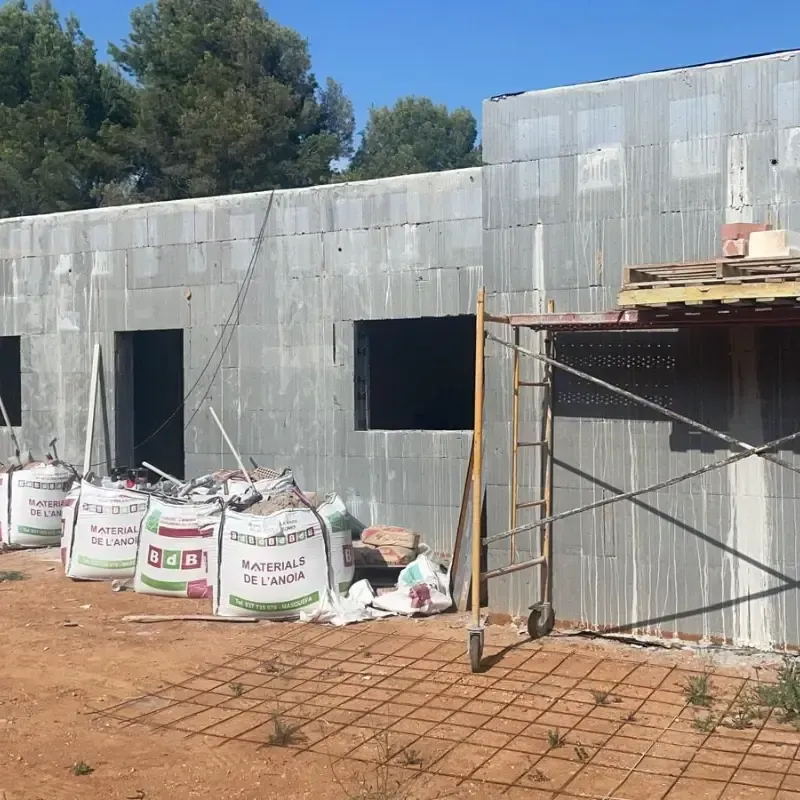 Edificio de chapa ondulada parcialmente construido, con ventanas abiertas, sacos de obra y andamios en el exterior.