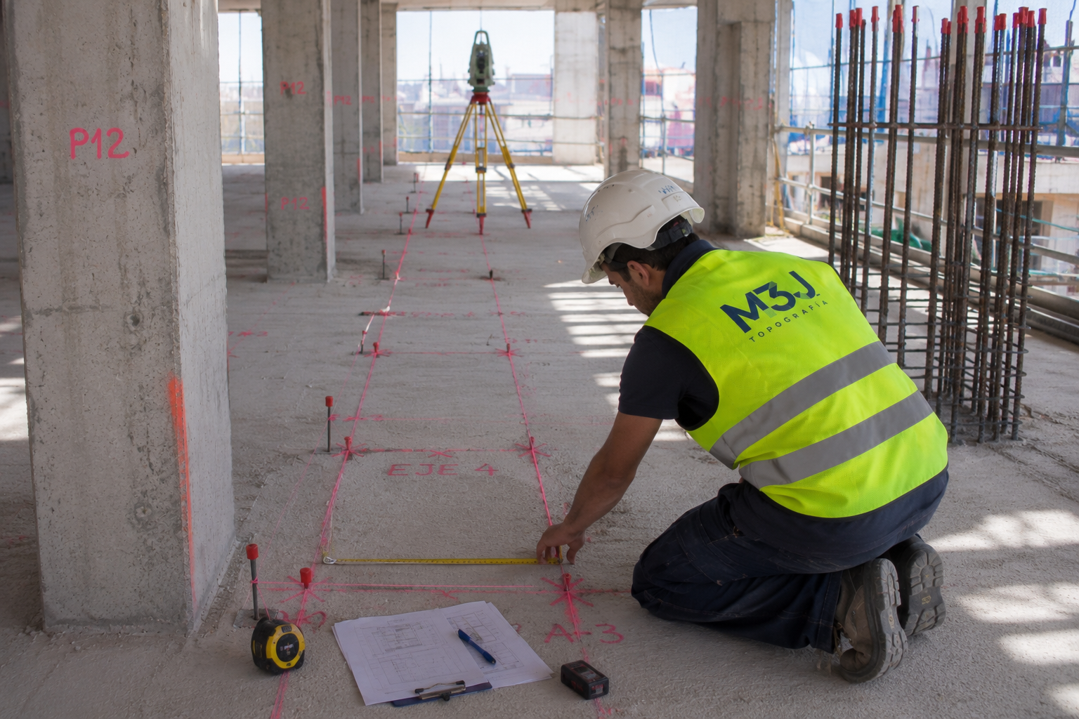 Un obrero de la construcción con chaleco amarillo se arrodilla midiendo un suelo de hormigón con marcadores rojos