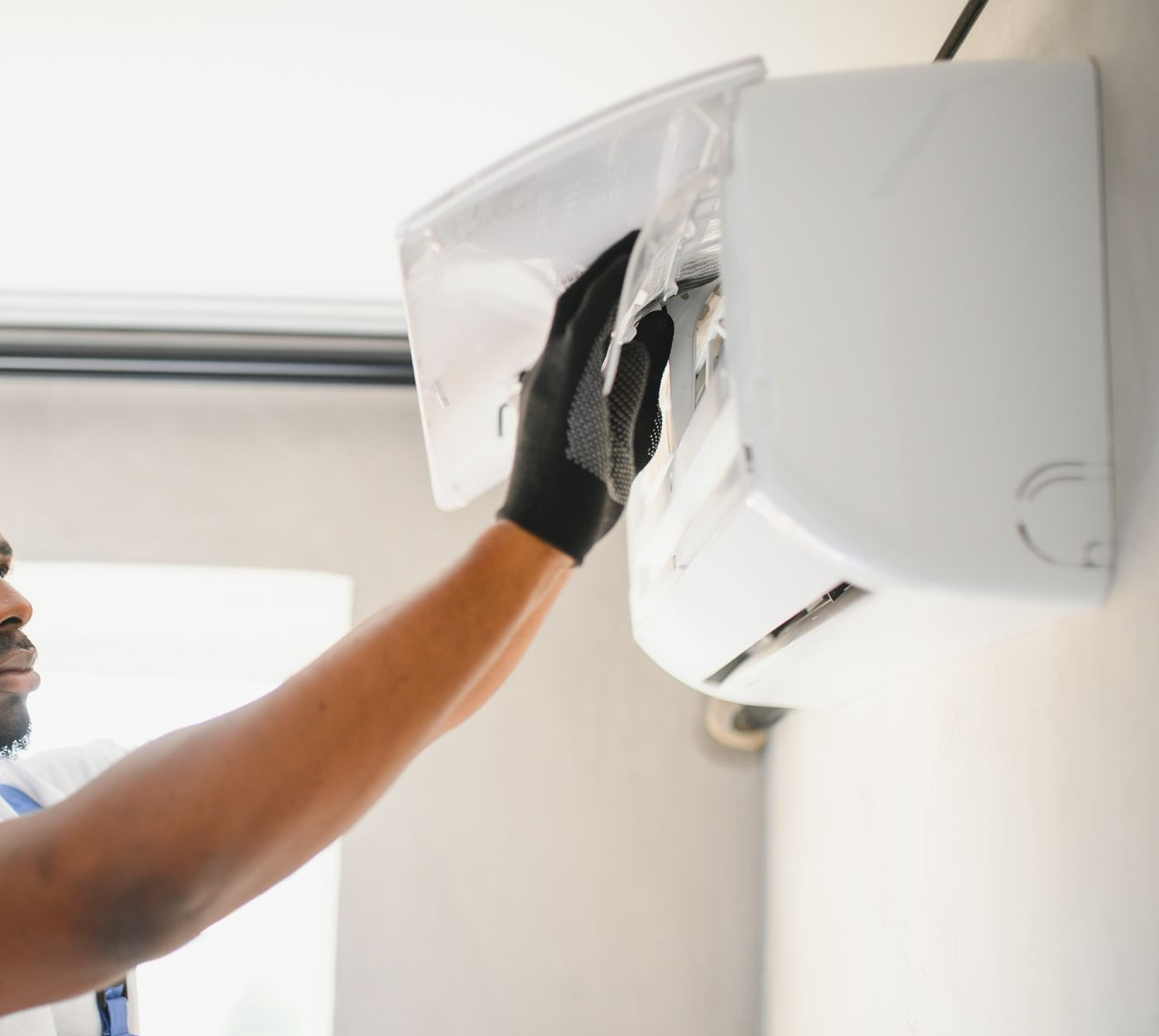 Un technicien portant des gants noirs nettoie l'intérieur d'un climatiseur mural blanc.