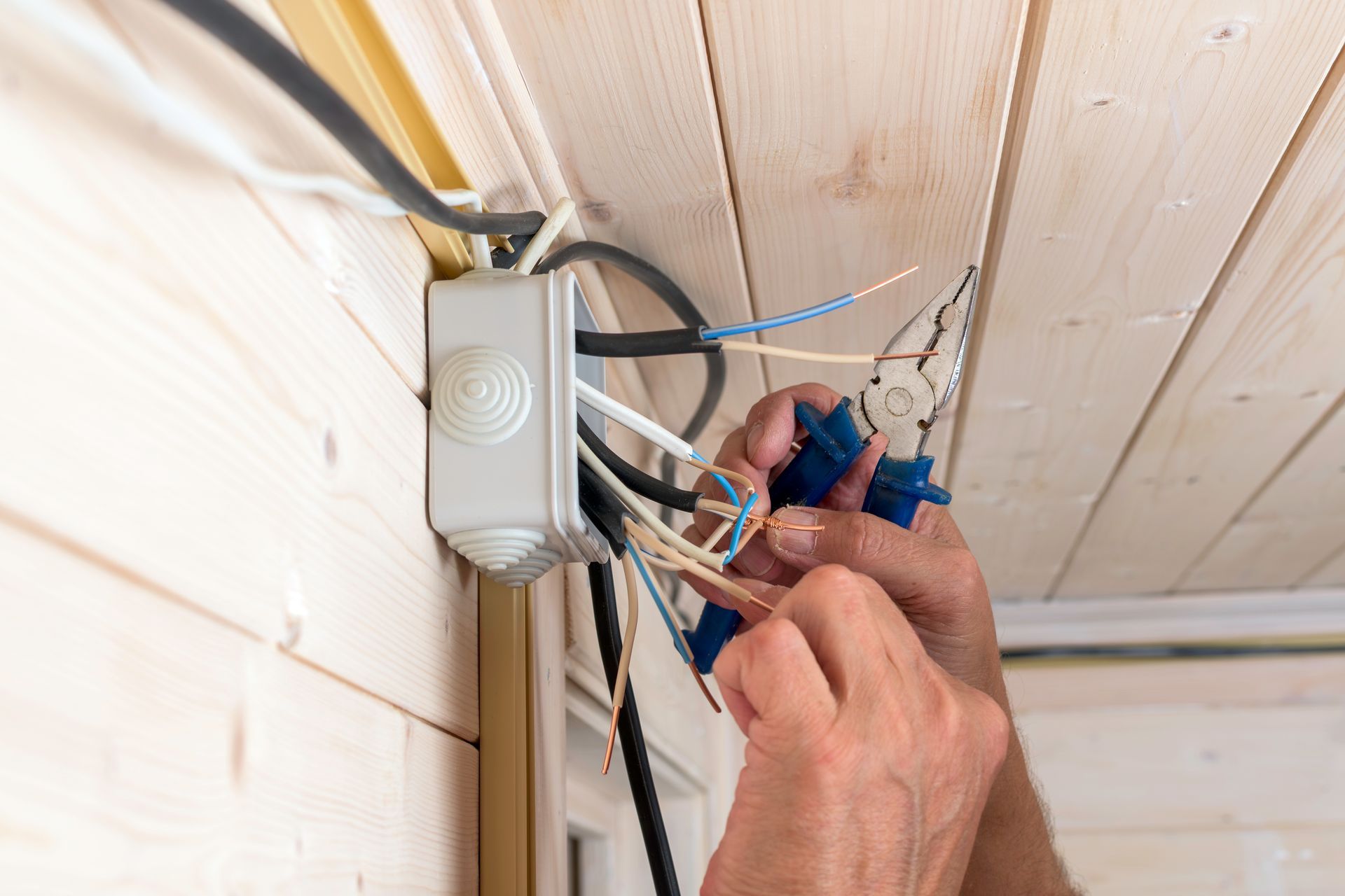 Une personne utilisant une pince pour câbler une boîte de jonction grise fixée à un mur en bois clair.