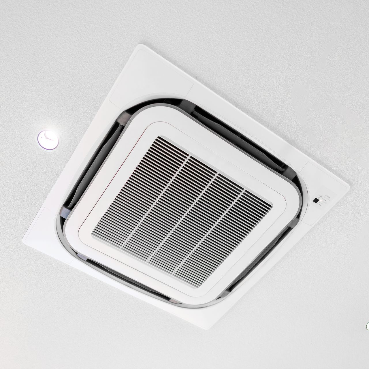 Unité de climatisation carrée blanche fixée au plafond, avec une grille d'entrée d'air central.