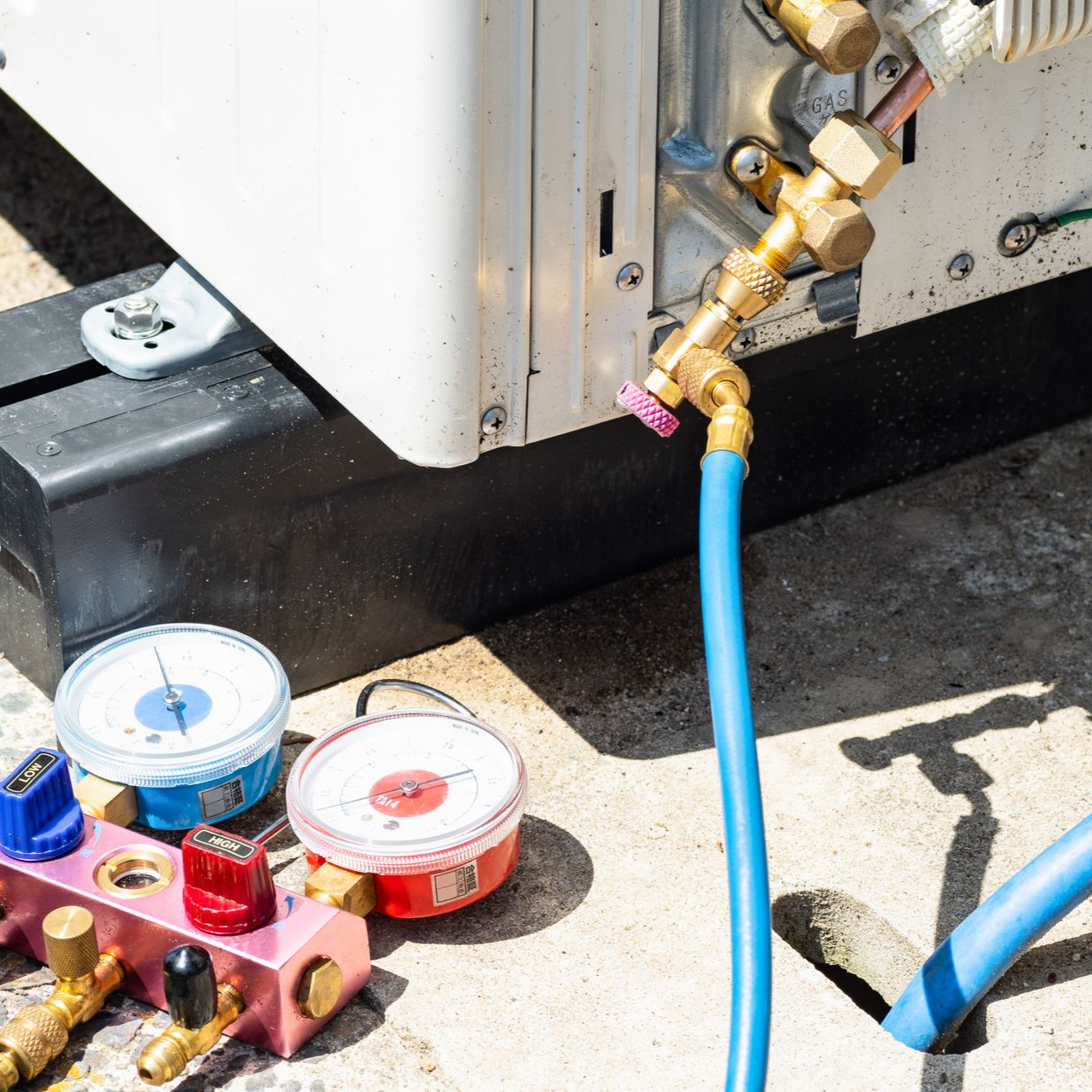 Outils pour technicien de CVC, notamment un manomètre relié par des tuyaux bleus à une unité de climatisation extérieure.
