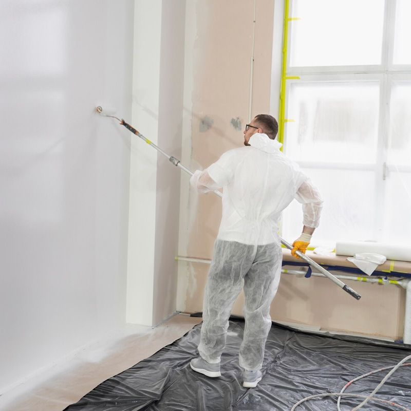 Persona con traje de protección pintando una pared blanca con un rodillo. En el interior, cubriendo el suelo.