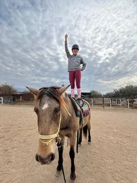 Una mujer está parada encima de un caballo en un campo de tierra.