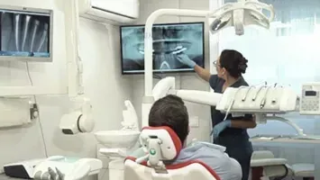 Un dentista y su asistente atienden a un paciente en una luminosa clínica dental, con monitores que muestran radiografías.