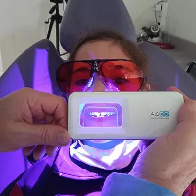Una persona sentada en un sillón dental, con gafas protectoras de color naranja, mientras se le realiza una prueba de fluorescencia oral.