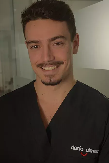 Persona sonriente con una blusa negra de pie en el interior de un edificio, con una pared clara de fondo.