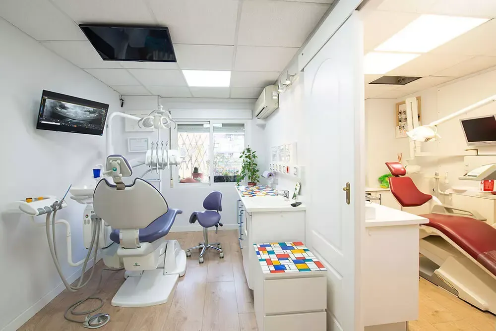 Clínica dental luminosa con dos sillones de tratamiento, equipo y gabinetes blancos bajo luces de techo.