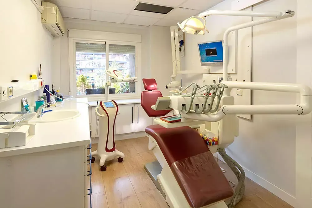 Sala de tratamiento dental con sillón rojo, armarios blancos y equipo suspendido cerca de una ventana.