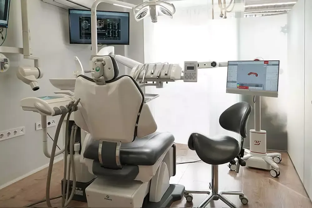 Clínica dental moderna con sillón de exploración, equipo y monitor en una habitación blanca y luminosa.