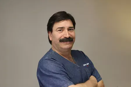 Hombre con uniforme médico azul y bigote, sonriendo con los brazos cruzados sobre un fondo beige liso.