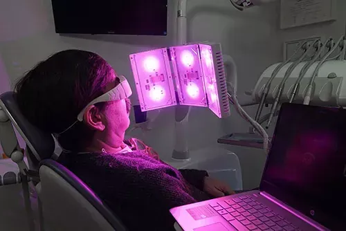 Una persona sentada en un sillón dental lleva gafas protectoras mientras mira un panel de luz LED violeta para fototerapia.