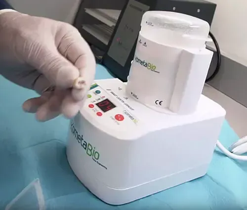 Una mano enguantada sostiene una corona dental junto a un horno de laboratorio dental 
