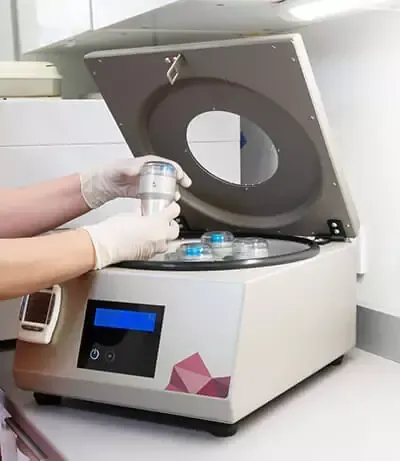 Una persona con guantes coloca un recipiente cilíndrico para muestras en una centrífuga de laboratorio abierta, situada sobre una encimera blanca.