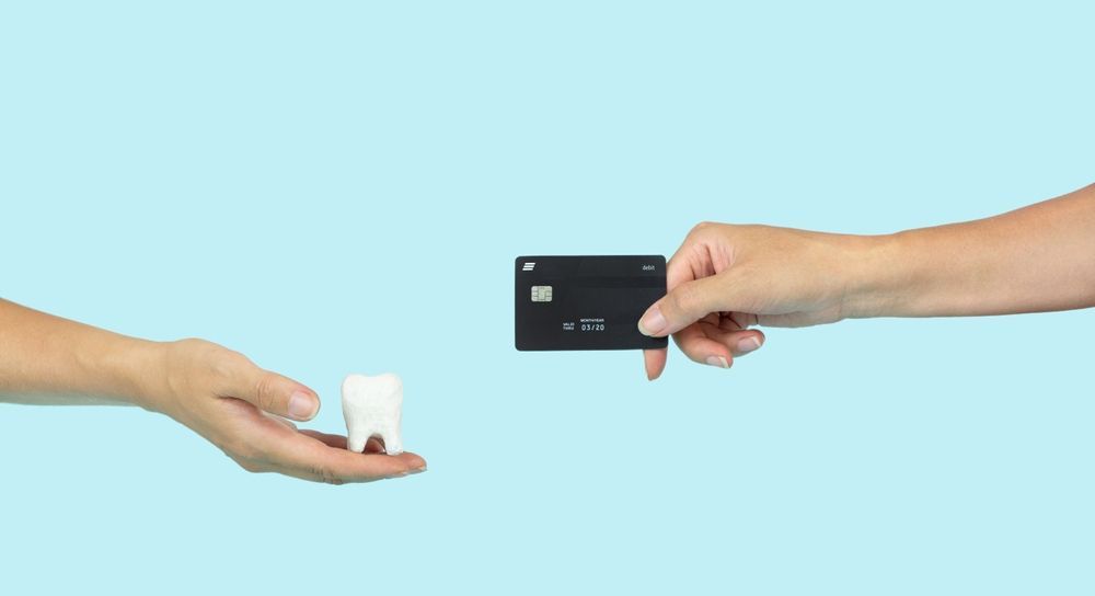 Dos manos intercambian una corona dental: una sostiene un pequeño diente blanco, la otra una tarjeta negra, sobre un fondo azul.