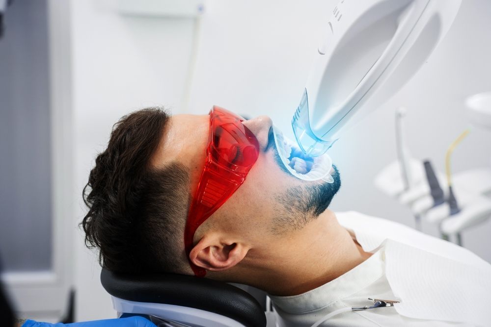 Una persona que lleva gafas protectoras rojas recibe un tratamiento profesional de blanqueamiento dental con un dispositivo de luz LED azul.