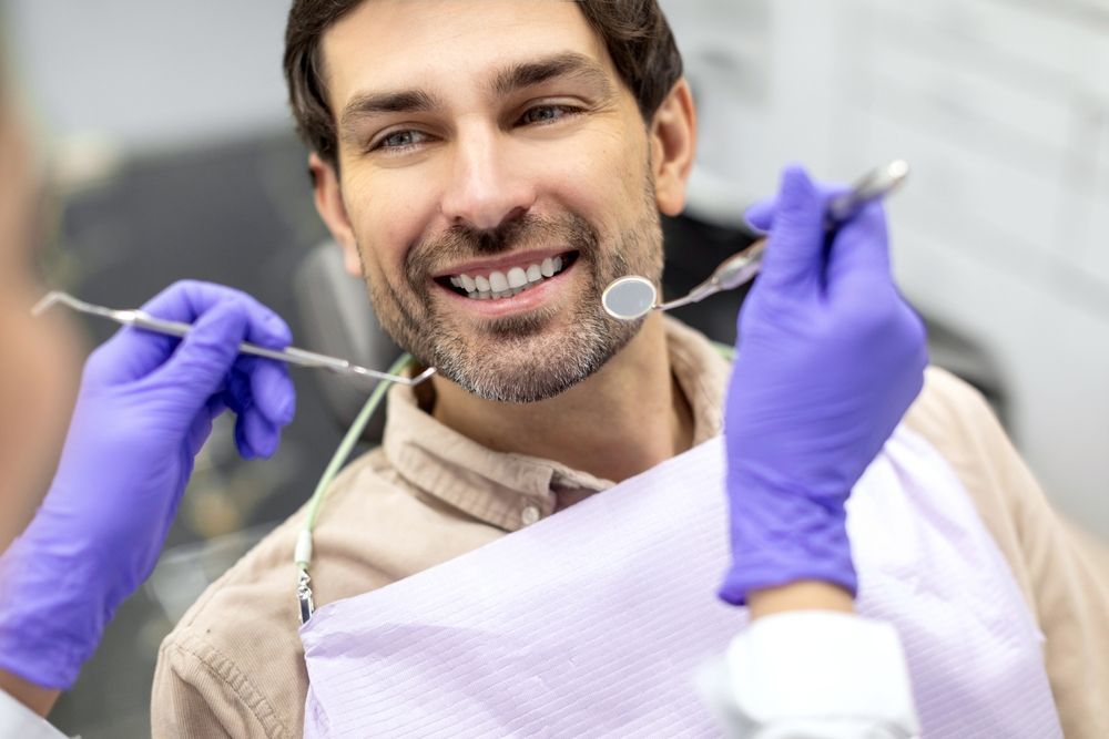 Un profesional dental con guantes morados utiliza un espejo y una sonda para examinar a un paciente sonriente sentado en el sillón dental.