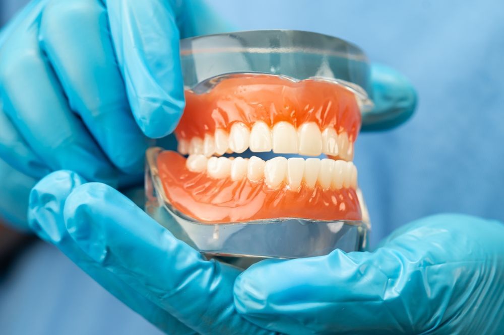 Primer plano de unas manos enguantadas que sostienen una dentadura postiza completa en un soporte transparente.