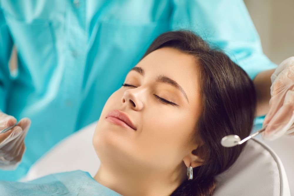 Un dentista con bata azul y guantes sostiene un espejo dental cerca de un paciente relajado que está recostado en un sillón dental.