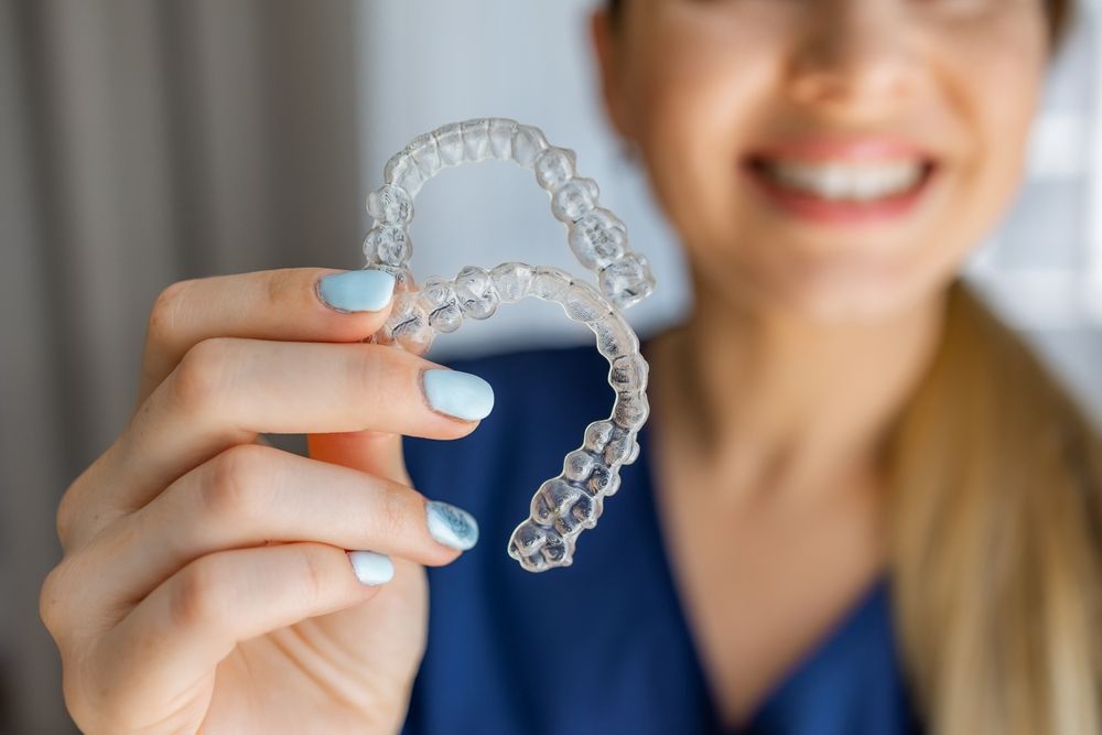 Una persona con una bata azul de uniforme médico sostiene una férula transparente para alineadores dentales sobre un fondo interior borroso.