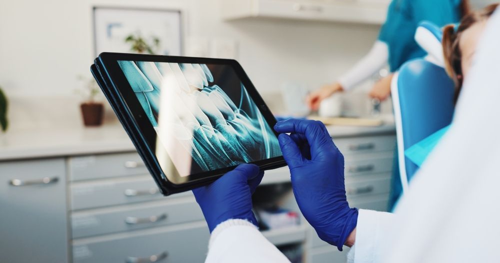 Un dentista con guantes azules sostiene una tableta que muestra una radiografía dental en un consultorio clínico.