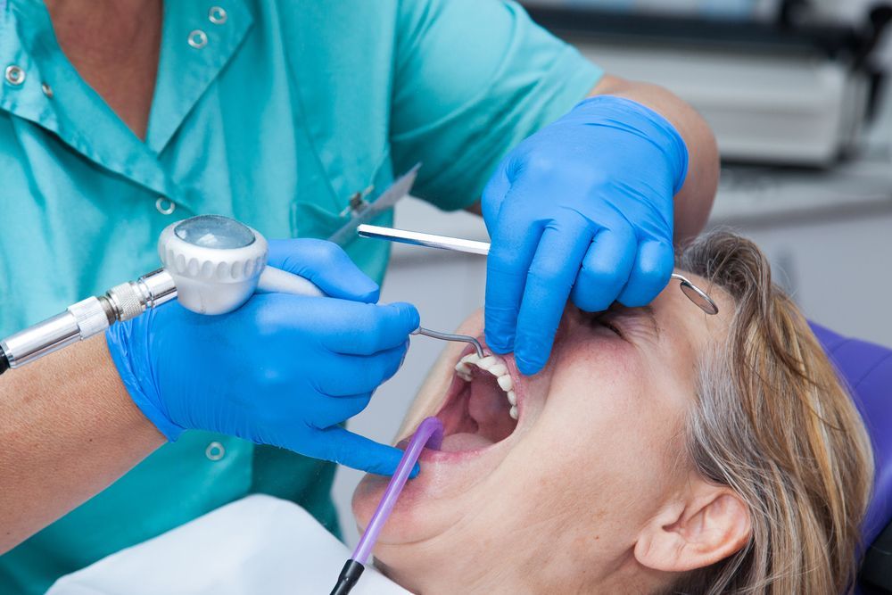 Un dentista, vestido con una bata quirúrgica de color turquesa y guantes azules, utiliza un instrumento dental para trabajar en los dientes de un paciente en un entorno clínico.