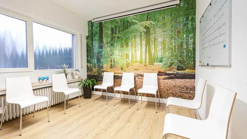 Ein Wartezimmer mit weißen Stühlen und einem Waldbild an der Wand.