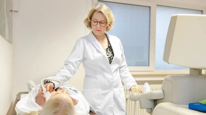 Eine Frau im weißen Kittel steht neben einem Patienten in einem Krankenhausbett.