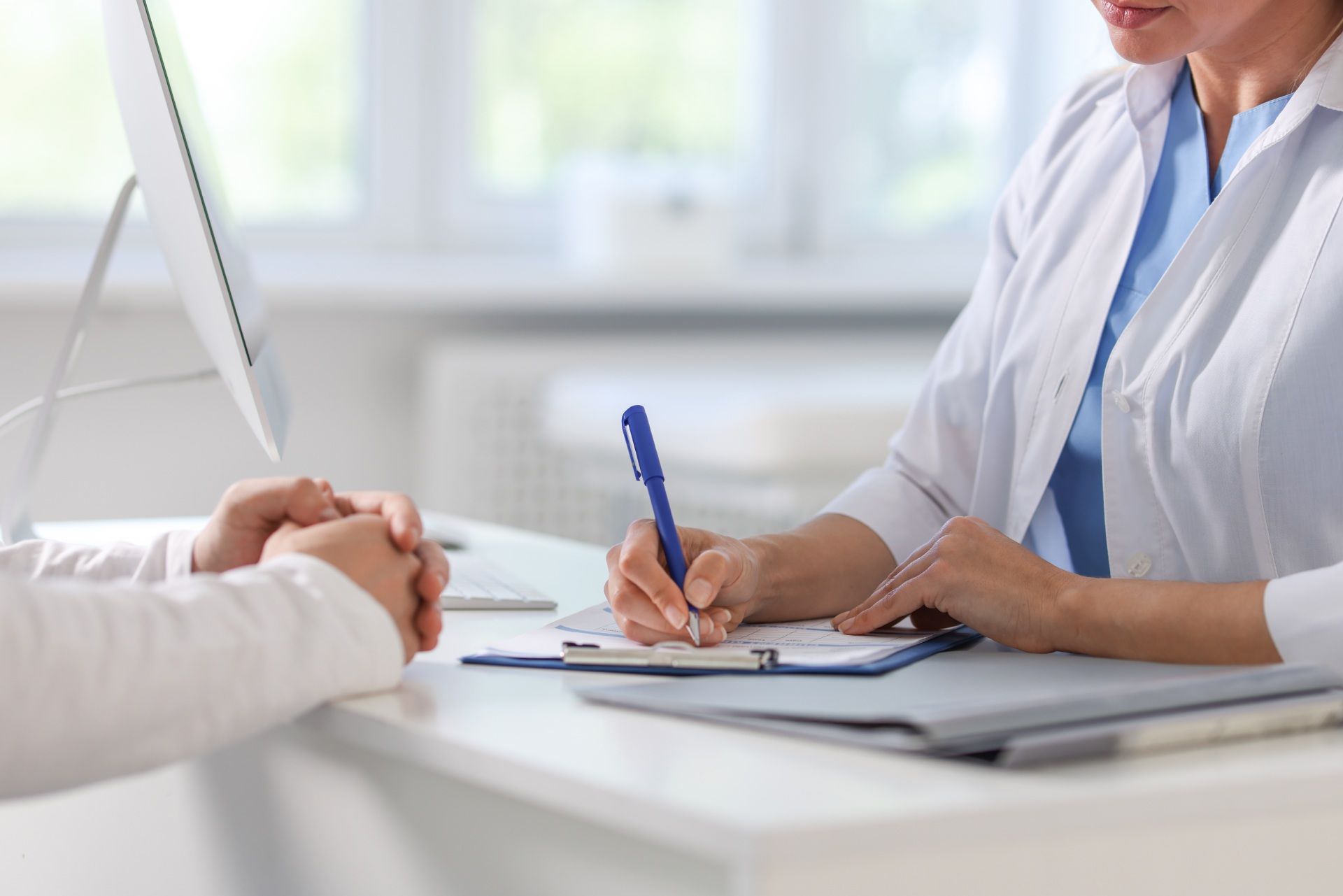 Ein Arzt sitzt mit einem Patienten an einem Schreibtisch und schreibt auf ein Klemmbrett.