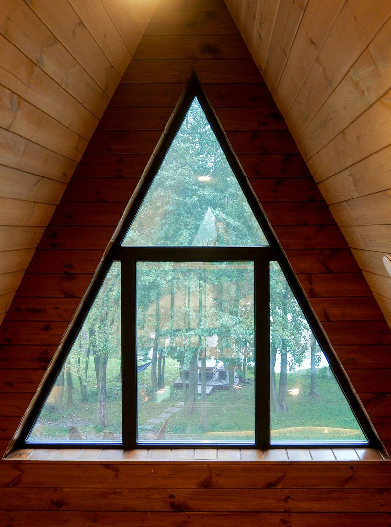 Fenêtre triangulaire dans une cabane, cadre foncé, vue sur les arbres et la verdure.