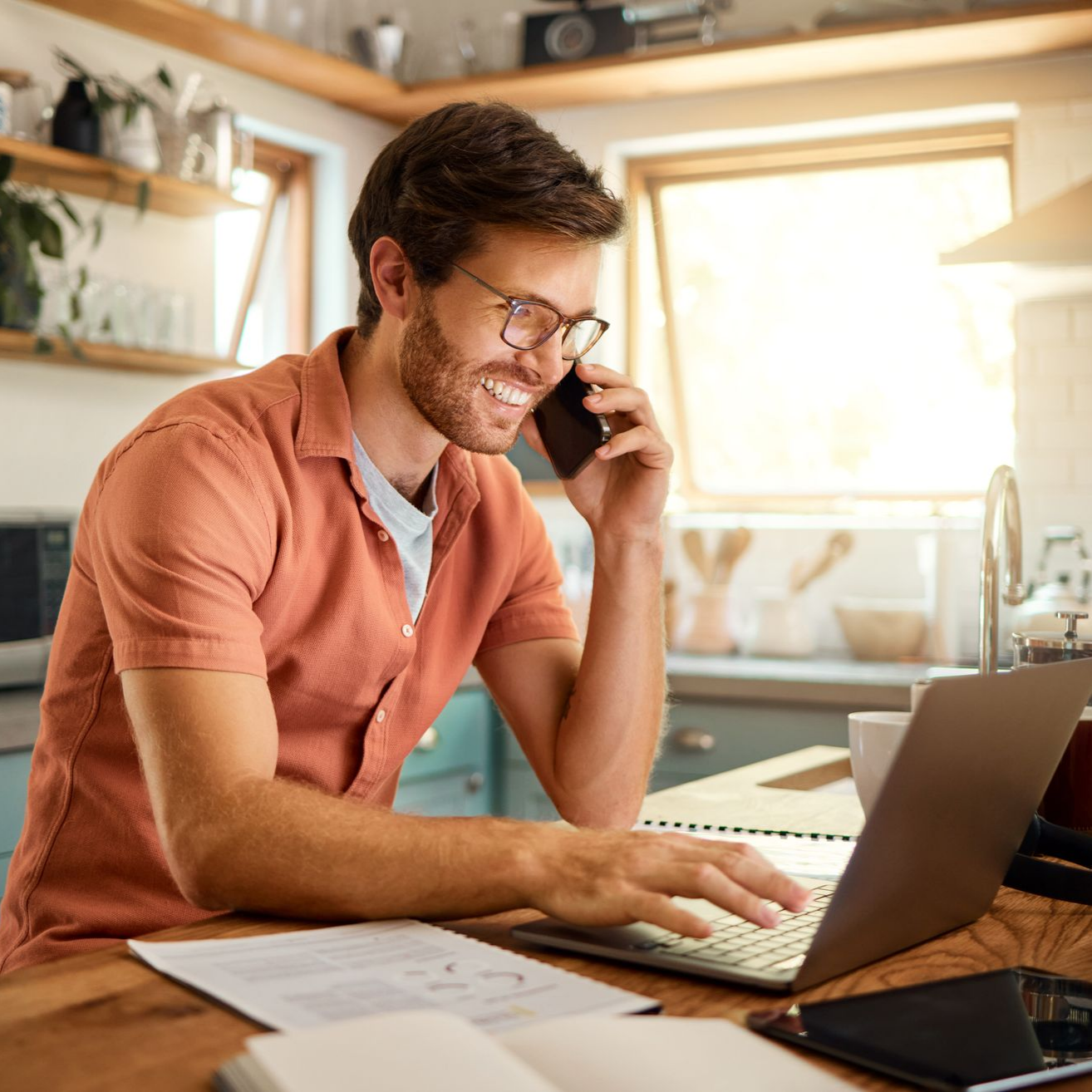 Un homme portant des lunettes au téléphone tout en travaillant sur son ordinateur portable dans sa cuisine.