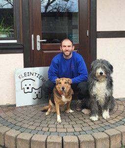 Jonas lächelt vor einem Praxiseingang, neben ihm sitzt ein wuscheliger, grau-weißer Hund.