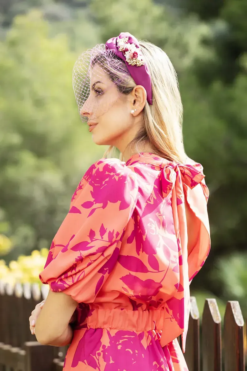 Una mujer con un vestido rosa y naranja y una diadema está parada frente a una valla de madera.