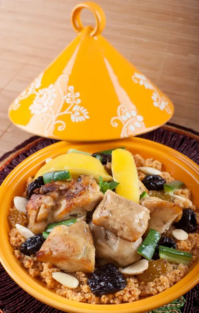 Plat à tajine orange avec du poulet, des légumes et des raisins secs, accompagné d’un couvercle jaune à motifs