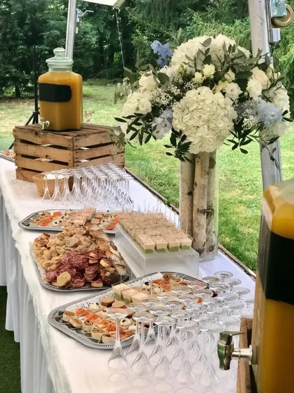 Table de buffet en plein air avec des amuse-bouches, des verres à vin et des compositions florales blanches sous une tente