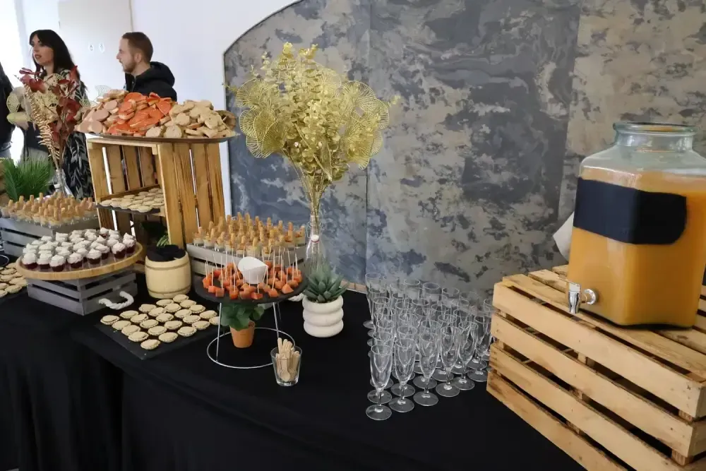 Buffet avec des viennoiseries, des fruits, des boissons et des gobelets empilés, près d’arbres.